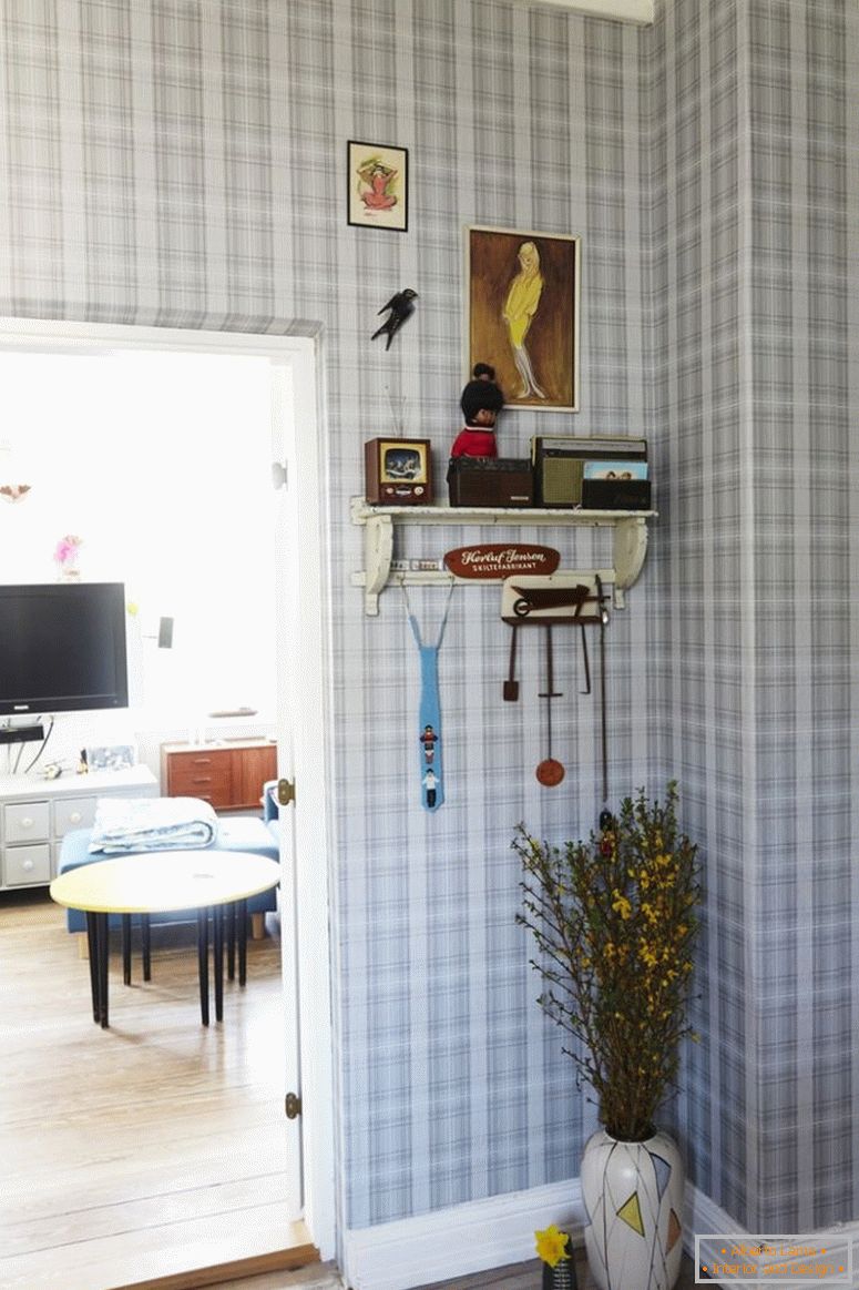 Pasillo con el papel pintado a cuadros, florero del piso debajo del tablero de la pared, al lado de la puerta abierta a la sala de estar