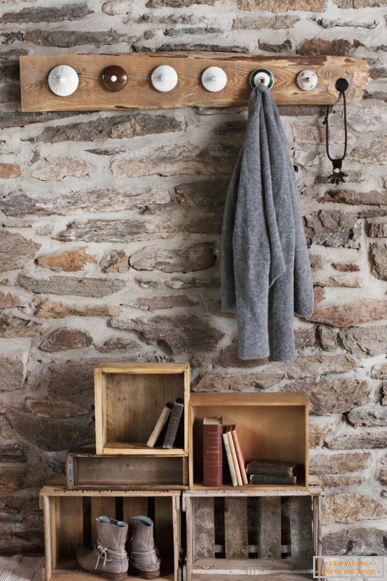 Rústico guardarropa de bricolaje en la habitación con muro de piedra; perchas hechas de tapas de porcelana vieja y cajas de madera viejas en el piso como almacenamiento